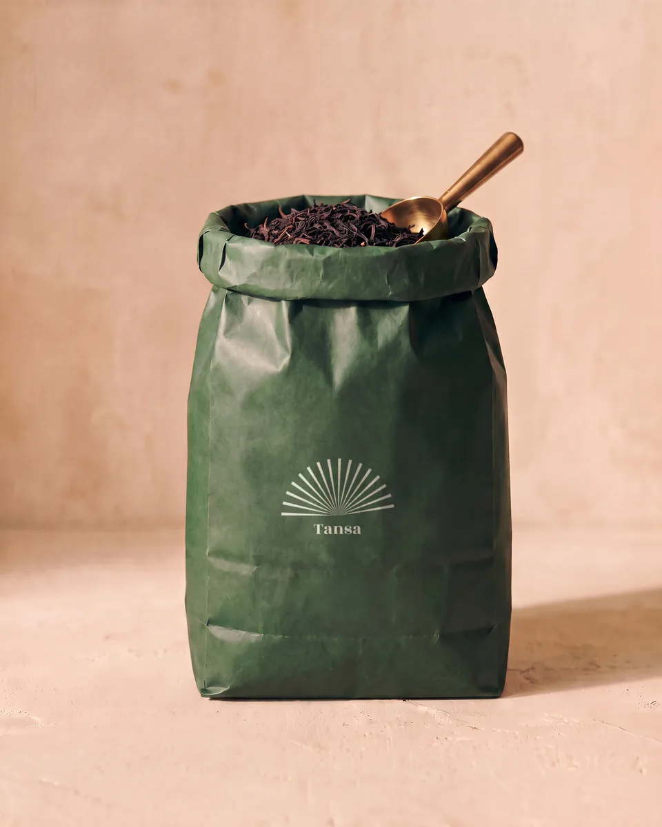 A large green paper bag of loose tea with a brass scoop and a cream sunburst.