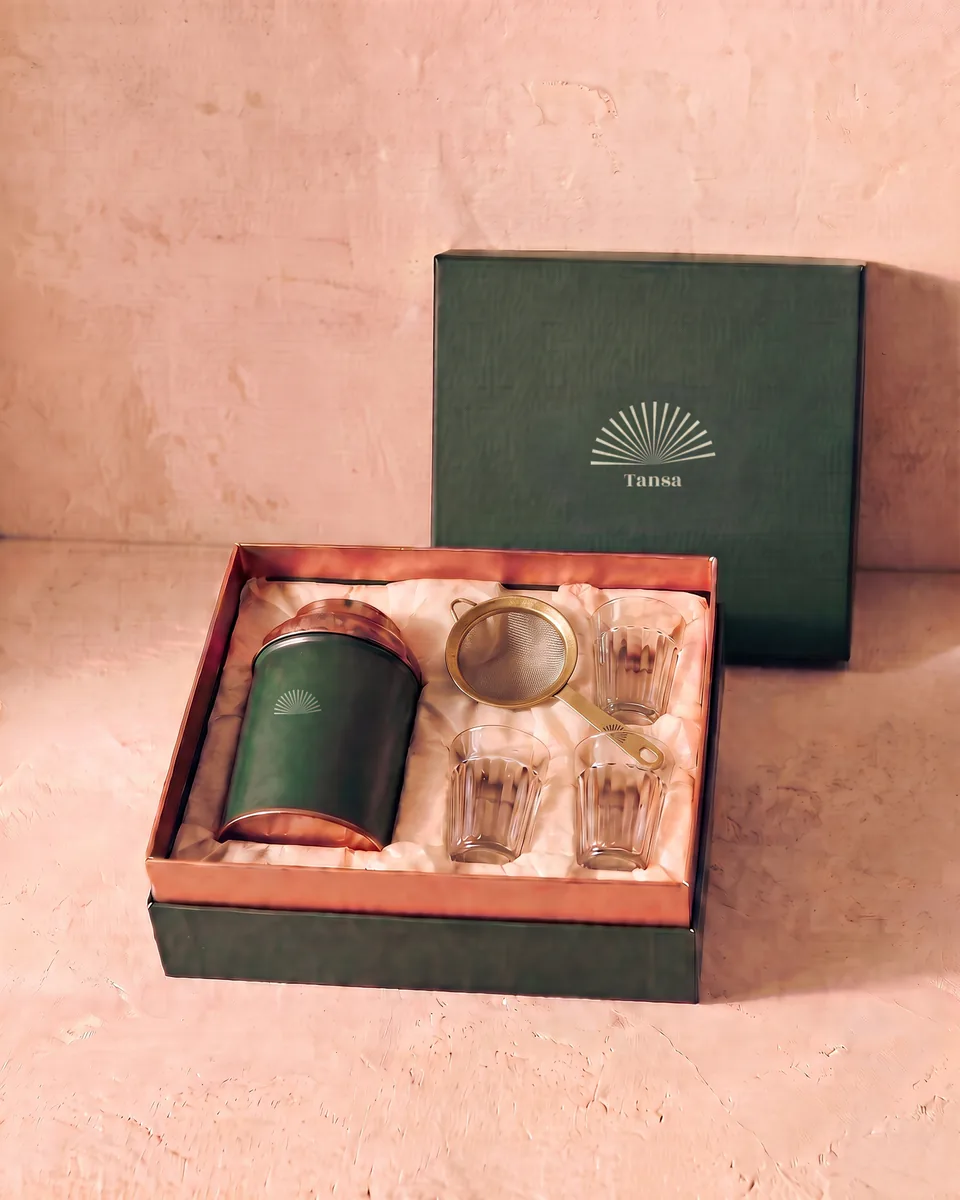 A green gift box with a sunburst, holding the house tin, a brass strainer and four chai glasses.
