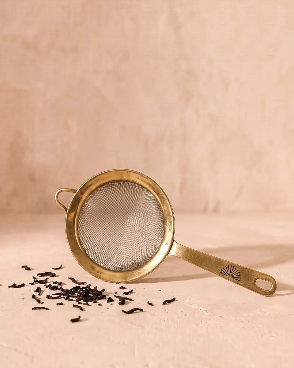 A brass-rimmed tea strainer with a long brass handle, a few tea leaves beside it.